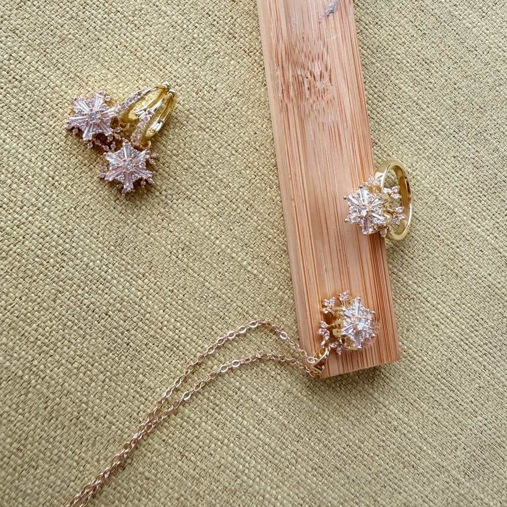 A gold jewelry organizer made of a bamboo wood block with three burnished holes, displayed on a beige linen background. The organizer is shown holding a gold ring, a gold necklace, and a pair of gold snowflake stud earrings.