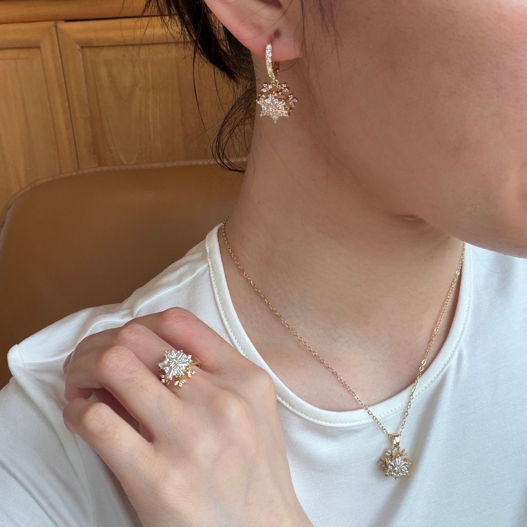 A close-up lifestyle shot of a person wearing a matching gold jewelry set, featuring a ring, necklace, and earring with a floral starburst design set with clear crystals.