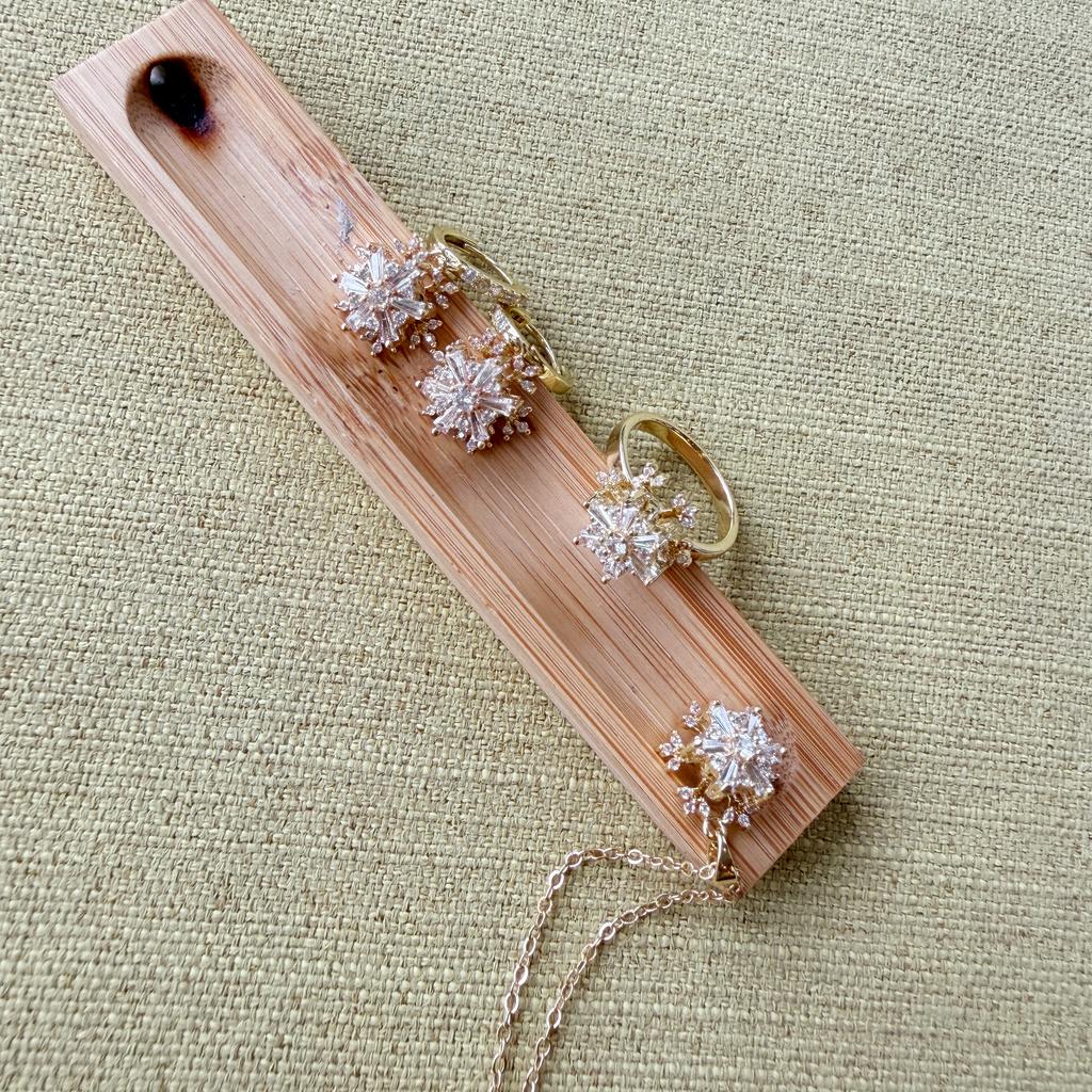 A set of gold-toned jewelry pieces displayed on a light wood block over a textured beige fabric background. The set includes three snowflake-shaped earrings and a matching snowflake ring with a delicate chain.