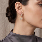 A close-up of a woman's ear and neck wearing a delicate gold huggie hoop earring with a dangling floral cluster of clear pavé stones.