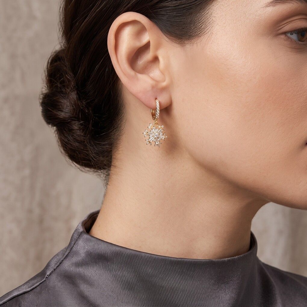 A close-up of a woman's ear and neck wearing a delicate gold huggie hoop earring with a dangling floral cluster of clear pavé stones.