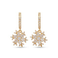 A pair of gold huggie hoop earrings with snowflake-shaped drop charms. The hoops are paved with small clear stones, and the snowflake charms feature baguette-cut clear stones arranged in a six-pointed star design, all set in yellow gold against a plain white background.