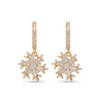 A pair of gold huggie hoop earrings with snowflake-shaped drop charms. The hoops are paved with small clear stones, and the snowflake charms feature baguette-cut clear stones arranged in a six-pointed star design, all set in yellow gold against a plain white background.