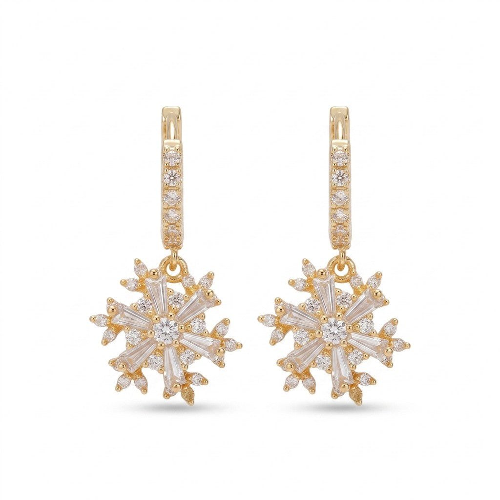 A pair of gold huggie hoop earrings with snowflake-shaped drop charms. The hoops are paved with small clear stones, and the snowflake charms feature baguette-cut clear stones arranged in a six-pointed star design, all set in yellow gold against a plain white background.