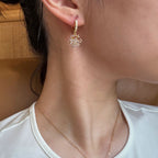 A close-up of a person's ear and neck wearing a delicate gold huggie hoop earring adorned with small clear crystals in a snowflake or starburst design, paired with a fine gold chain necklace.