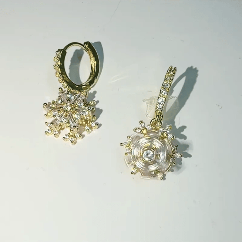 A pair of gold hoop earrings with dangling snowflake and spiral flower charms, embellished with clear rhinestones, shown on a plain white surface.