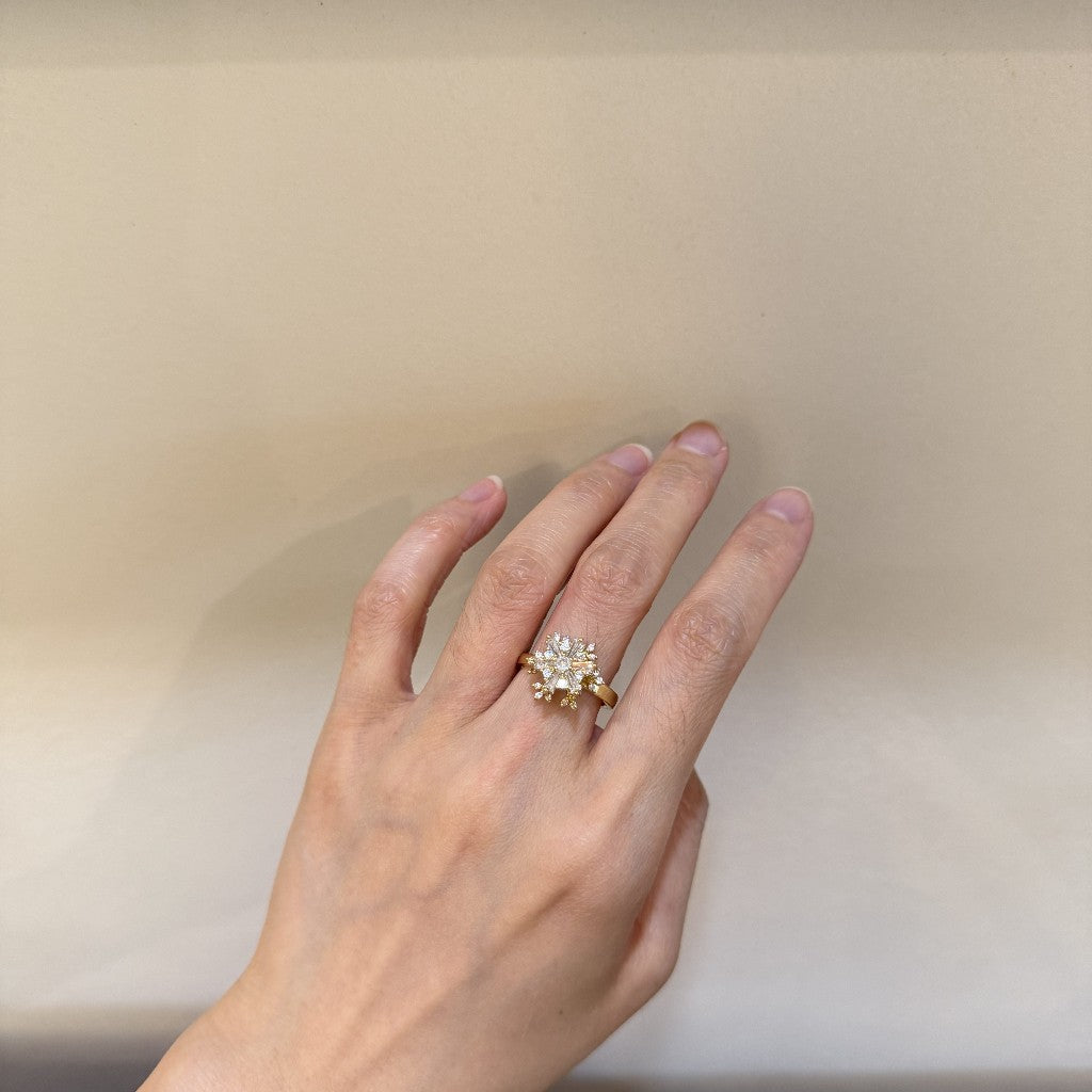 A gold ring with a clear crystal sunburst design worn on a person's ring finger, shown against a plain beige background.