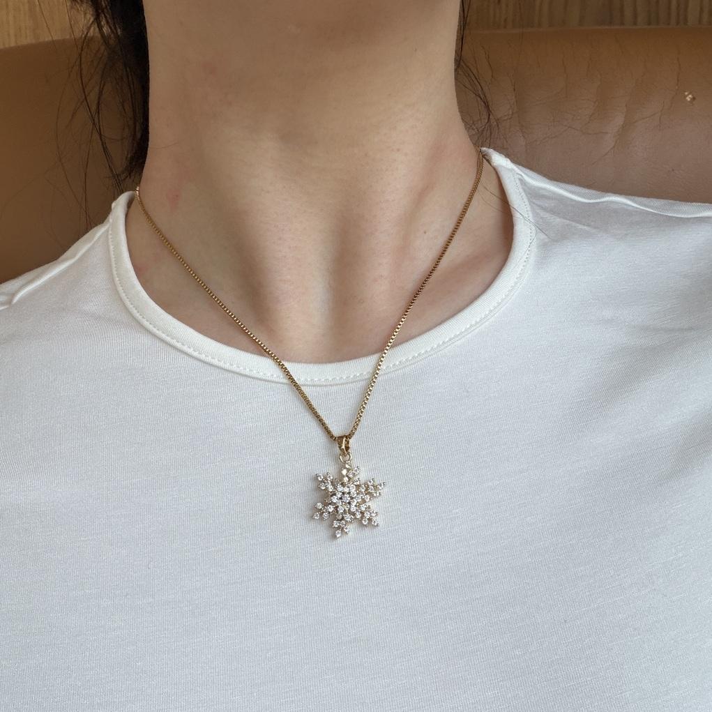 A close-up of a person's neck and chest wearing a delicate gold chain necklace with a snowflake-shaped pendant adorned with small clear rhinestones, paired with a white crew-neck t-shirt.