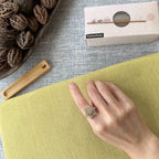A hand wearing a silver-toned snowflake-shaped ring with clear stones, resting on a yellow-green textured surface. A white box labeled FushanKodo is visible in the background along with a wooden tool and walnuts on a gray fabric surface.