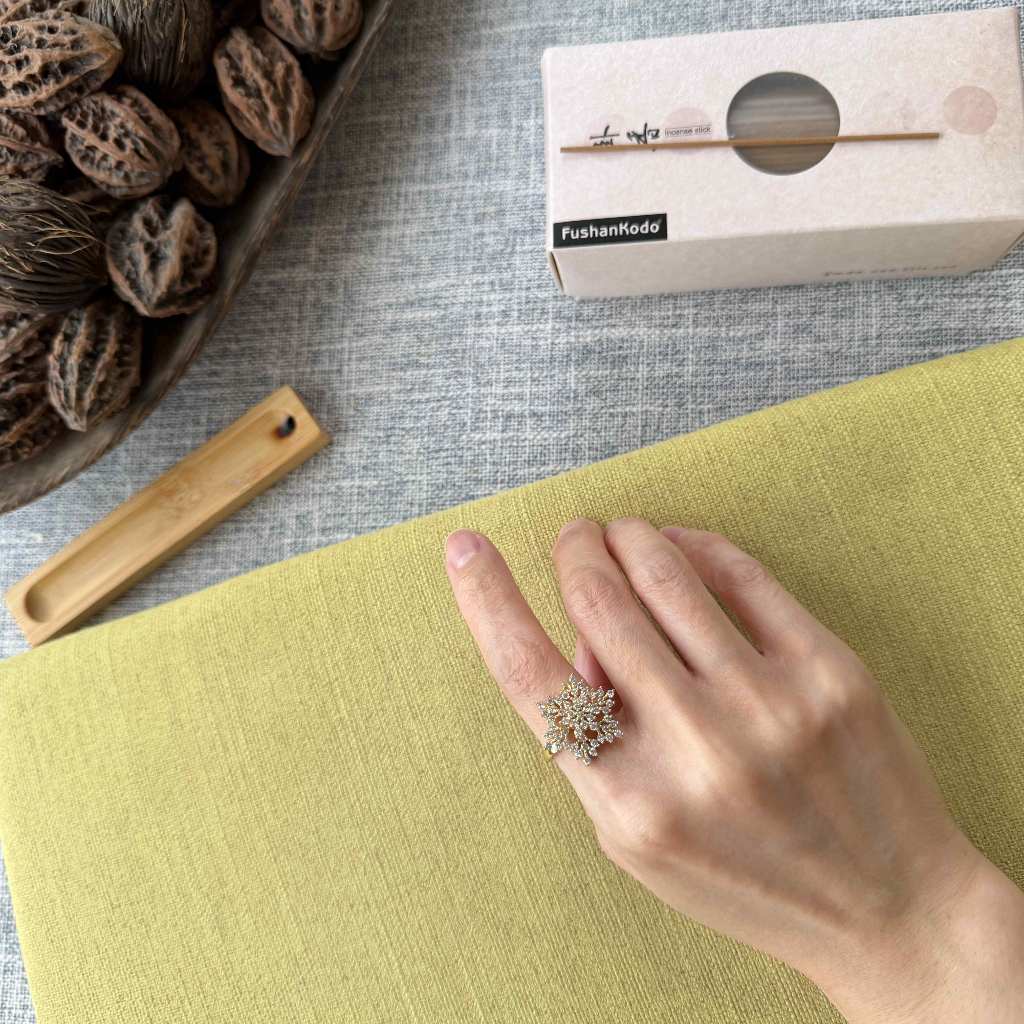 A hand wearing a silver-toned snowflake-shaped ring with clear stones, resting on a yellow-green textured surface. A white box labeled FushanKodo is visible in the background along with a wooden tool and walnuts on a gray fabric surface.