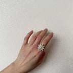 A silver-toned starburst ring with clear crystals worn on a person's hand, set against a textured off-white wall.