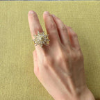A gold ring with a large, starburst-shaped cluster of clear gemstones worn on a person's finger, set against a textured olive green background.