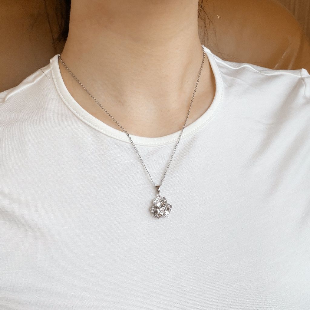 A close-up of a person wearing a silver chain necklace with a clear rhinestone flower-shaped pendant, styled with a white crew-neck t-shirt.