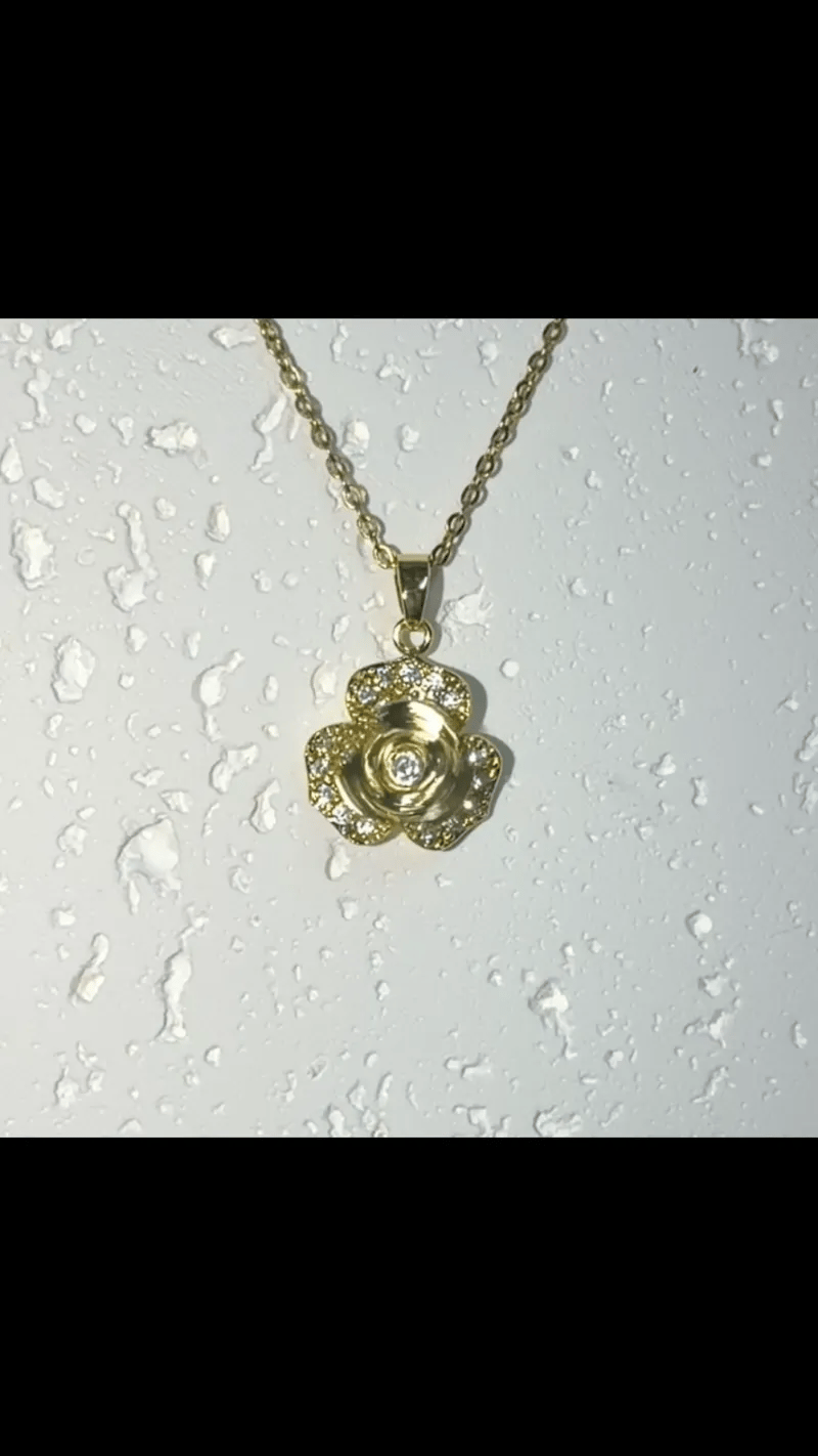 A gold-tone rose-shaped pendant necklace with a matching chain, displayed against a white textured surface with water droplets.