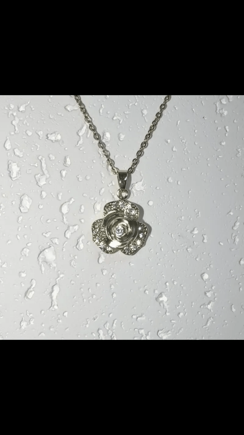 A silver-tone flower pendant necklace with a clear central gemstone, hanging from a matching chain against a white background with water droplets.