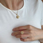 A close-up of a person wearing a gold flower-shaped pendant necklace and a matching gold flower ring on their finger, styled with a white crew-neck t-shirt.