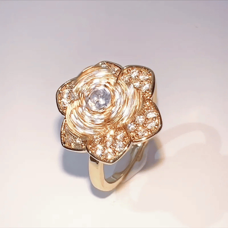 A gold flower-shaped ring with a central clear gemstone and smaller pavé-set stones, photographed on a plain light background.