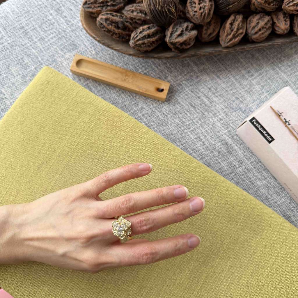 A hand wearing a gold flower-shaped ring with clear stones, resting on a textured yellow-green fabric surface. A wooden bowl of walnuts, a bamboo tool, and a small box labeled 'L'ART DES JOEUX' are arranged around it on a gray fabric background.