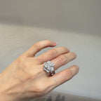 A silver-toned flower cluster ring with clear gemstones worn on a person's hand, set against a neutral gray wall.