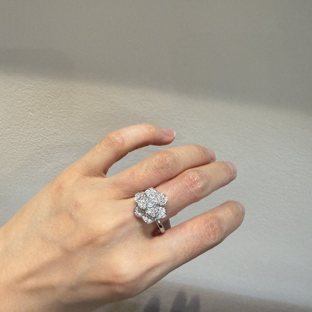 A silver-toned flower cluster ring with clear gemstones worn on a person's hand, set against a neutral gray wall.