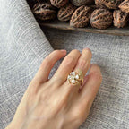 A hand wearing a gold ring with a floral cluster of clear stones, resting on a textured gray fabric next to a wooden tray filled with brown walnuts.