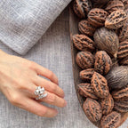 A silver ring with a floral cluster design is worn on a person's hand, which is resting on a textured gray linen cloth next to a wooden bowl filled with brown walnuts.