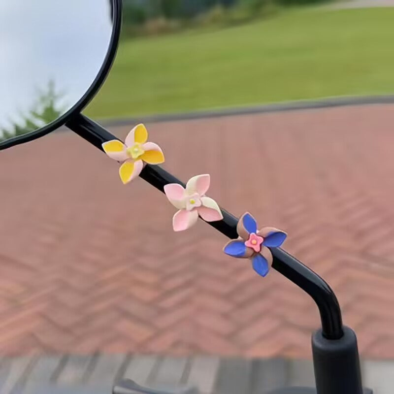 Three small flower-shaped decorative accessories in yellow, pink, and blue are attached to a black handlebar, with a blurred outdoor background showing a brick roof and green grass.