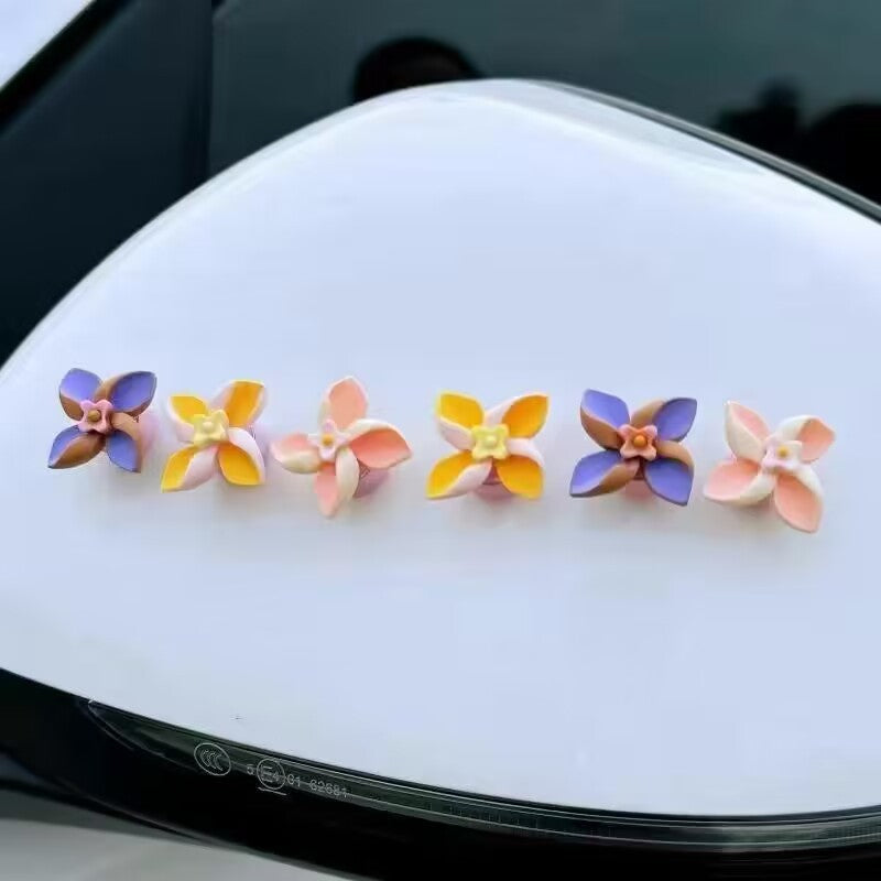 Five small flower-shaped accessories in assorted pastel colors arranged on a glossy white car mirror.