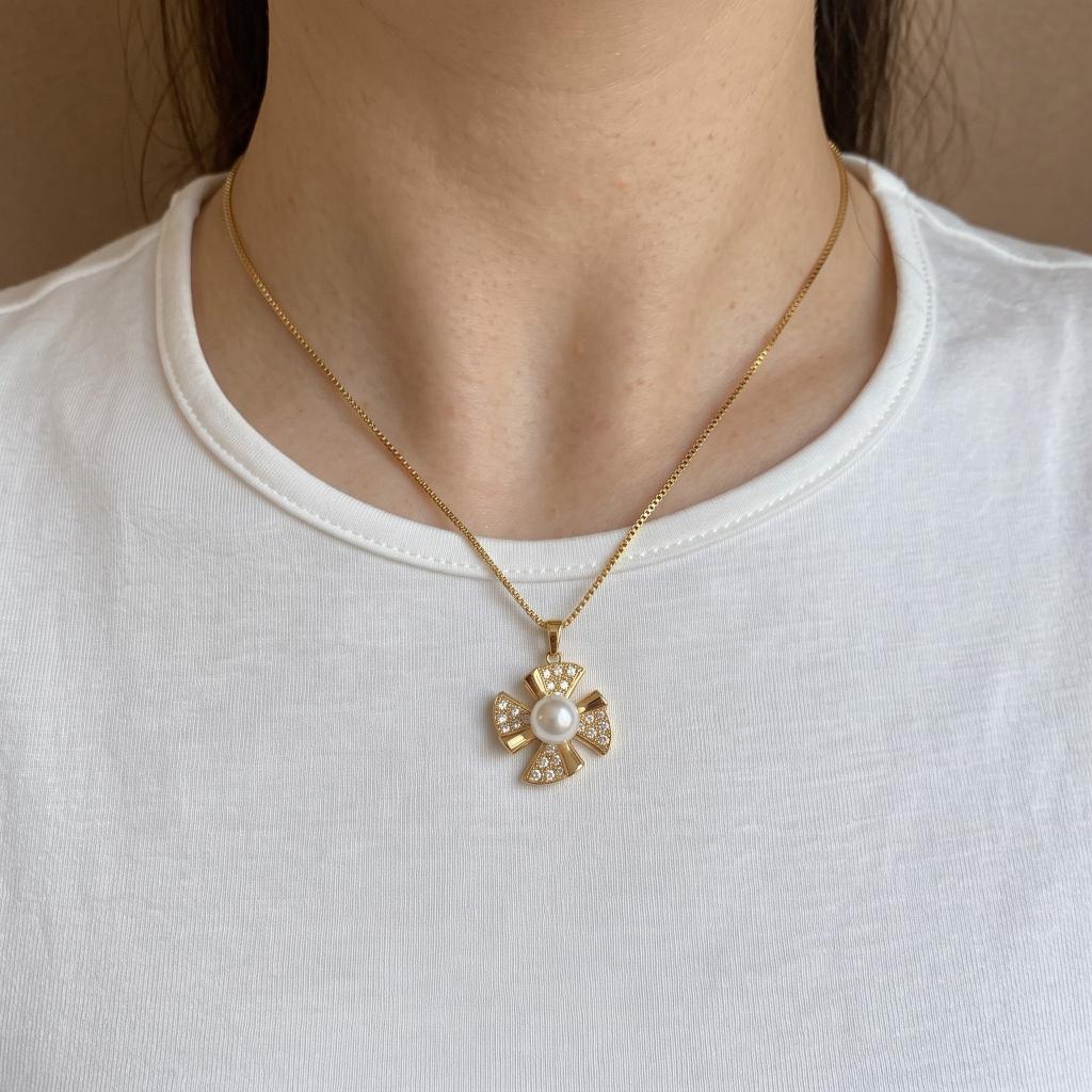 A gold cross pendant necklace with a central white pearl and small clear stones, worn on a model's neck over a white t-shirt.