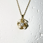 A gold-tone chain necklace with a spiral pendant featuring a white pearl in the center, displayed against a white textured background with water droplets.