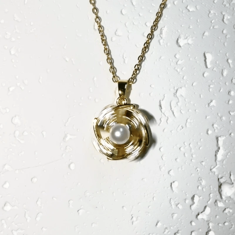 A gold-tone chain necklace with a spiral pendant featuring a white pearl in the center, displayed against a white textured background with water droplets.