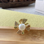 A gold flower-shaped ring with a central white pearl and small clear stones, resting on a wooden surface with a yellow-green textured mat and a blurred background.