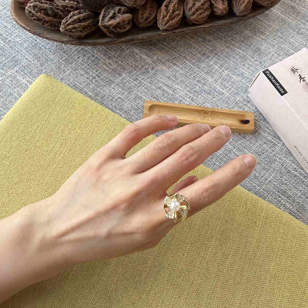 A close-up of a hand wearing a gold ring with a large clear floral-shaped gemstone, resting on a textured yellow-green mat. In the background, a wooden bowl filled with walnuts sits on a gray fabric surface next to a small wooden holder and a white box.