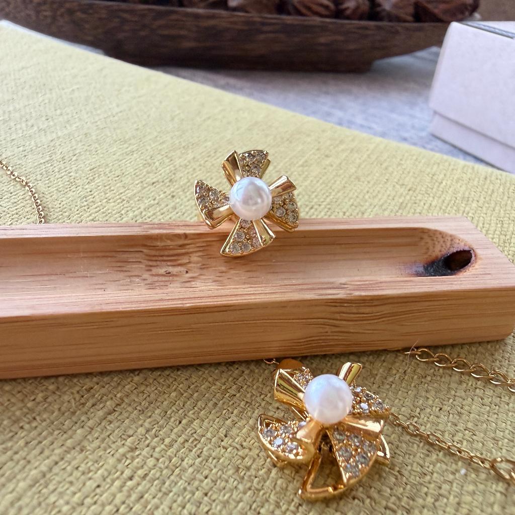 Gold-tone jewelry set featuring a flower-shaped pendant necklace and matching ring, both adorned with a central white pearl and small clear stones, displayed on a bamboo tray over a textured yellow-green fabric.