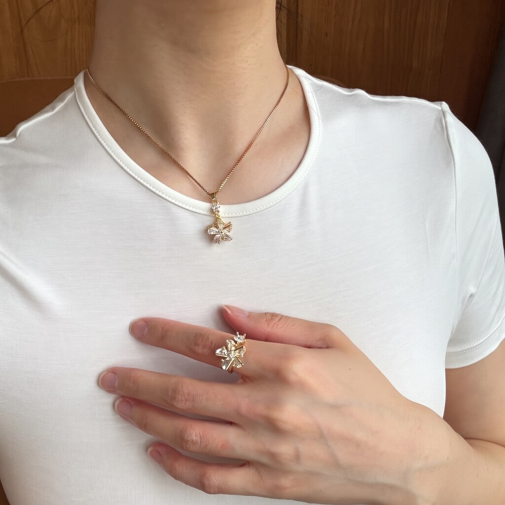 A close-up of a person wearing a delicate gold necklace with a small flower-like pendant and a matching gold ring on their finger, styled with a white t-shirt.