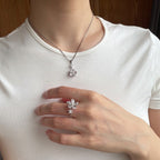 A person wearing a silver-toned flower-shaped pendant necklace and a matching flower ring on their finger, shown from the neck to the waist against a white t-shirt.