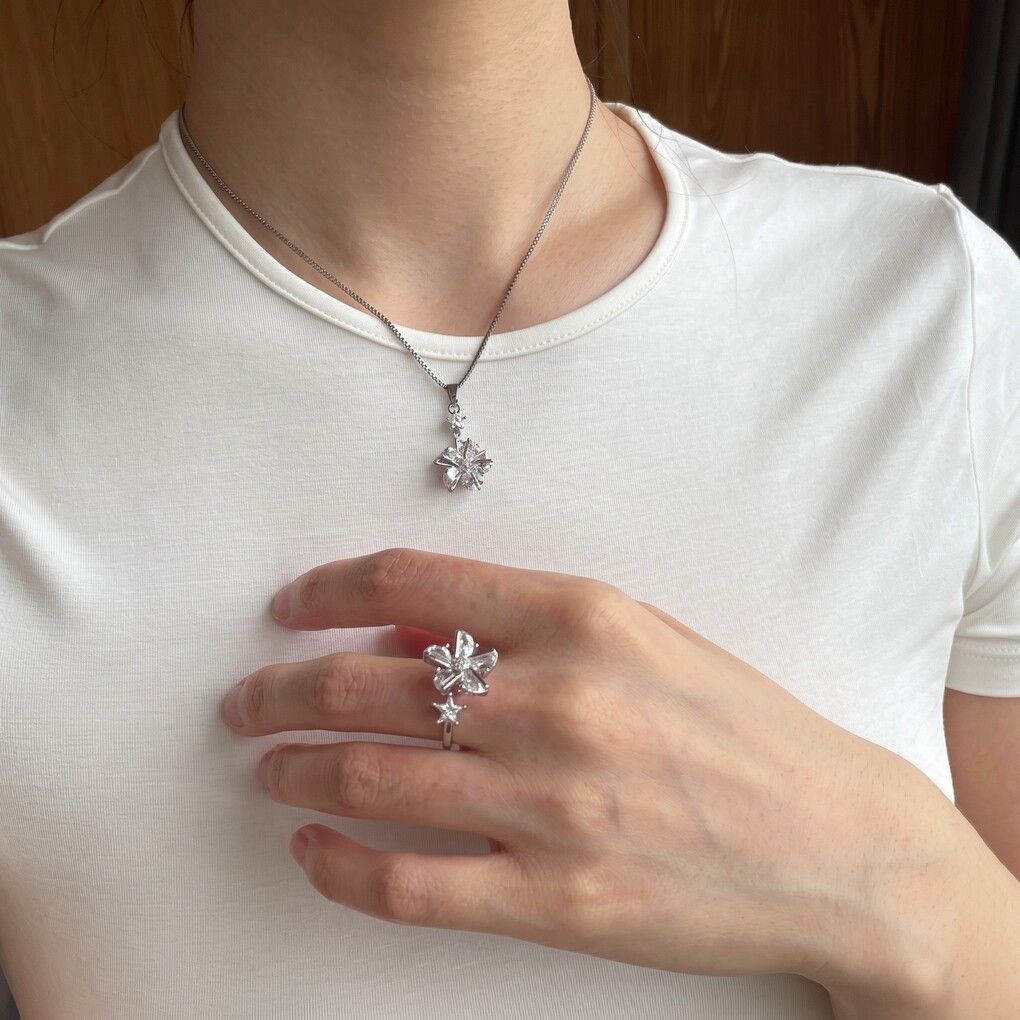 A person wearing a silver-toned flower-shaped pendant necklace and a matching flower ring on their finger, shown from the neck to the waist against a white t-shirt.