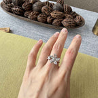 A silver-toned flower-shaped ring with clear stones worn on a person's hand, set against a background of a green cushion, textured gray tablecloth, and a wooden bowl filled with walnuts.