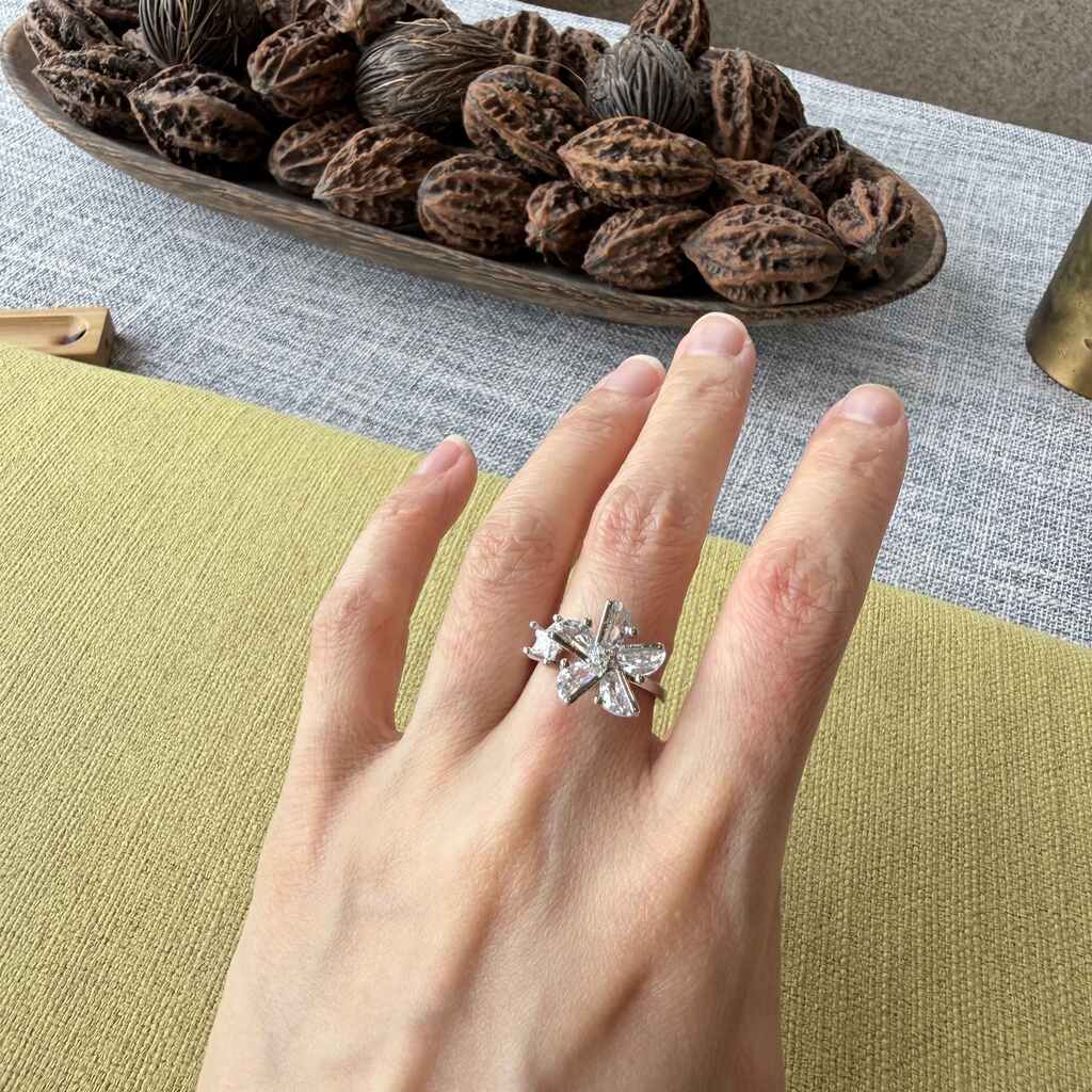 A silver-toned flower-shaped ring with clear stones worn on a person's hand, set against a background of a green cushion, textured gray tablecloth, and a wooden bowl filled with walnuts.