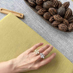A hand wearing two silver flower-shaped rings, resting on a textured olive-green surface with a gray woven mat and a wooden tray of walnuts in the background.