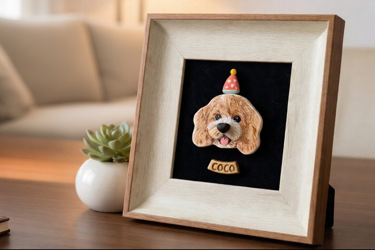 A custom pet portrait framed in a light wood square frame, featuring a 3D dog head sculpture with a party hat and a name plate reading 'COCO' on a black background, displayed on a wooden table next to a small potted succulent.