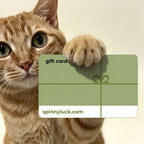 An orange tabby cat holds up a green gift card with its front paw, against a plain light beige background. The card reads 'gift card' and 'spinnyluck.